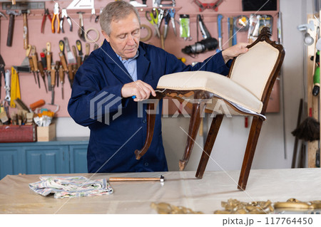 Skilled old furniture repair specialist in uniform restoring Victorian chair during workday in workshop 117764450