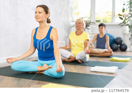 Middle-aged woman practicing half lotus pose of yoga in light fitness room Middle-aged woman practicing half lotus pose of yoga in light fitness room 117764579