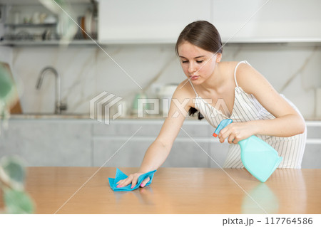 Positive young girl cleaning the surface of kitchen table Positive young girl cleaning the surface of kitchen table 117764586