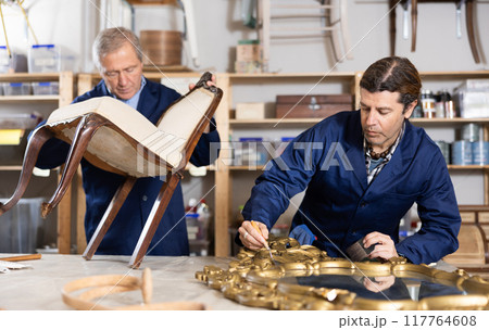Mature male carpenter and his apprentice working on restoring vintage mirror and chair in workshop 117764608