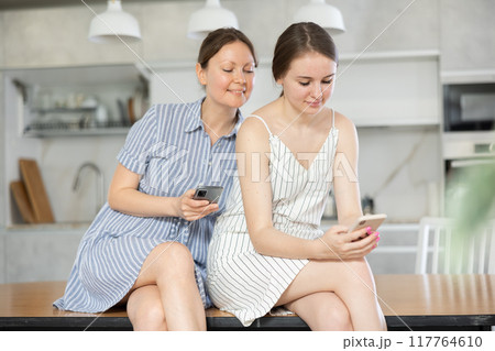 Mother and teen daughter looking at their phones sitting on kitchen-table 117764610