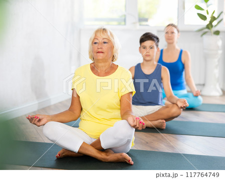 Old woman practicing half lotus pose of yoga in light fitness room 117764749