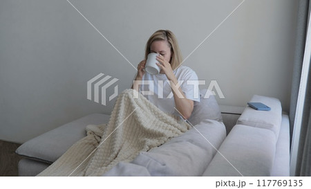 Young woman covered with a warm blanket drinks tea at home. A young woman drinks hot tea while lying on the sofa. A woman on the sofa, covered with a blanket, drinks a hot drink. 117769135