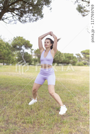 A Young Woman Energetically Performing a Jumping Jack Exercise in a Lush Green Park on a Sunny Day A Young Woman Energetically Performing a Jumping Jack Exercise in a Lush Green Park on a Sunny Day 117769769