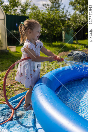 Closing the hose with his finger, the child sprays water, filling the pool. Splashes of water fly in all directions while the child plays with the hose, filling the pool. Summer concept. 117771942