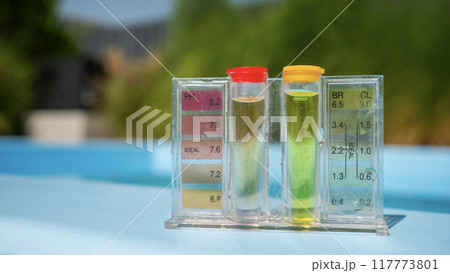 A pool water testing kit with pH and chlorine levels displayed in tubes, set against a blurred outdoor background. 117773801