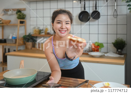 Cheerful sportswoman leaning on kitchen counter and eating toasted bread Cheerful sportswoman leaning on kitchen counter and eating toasted bread 117774712