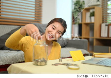 Happy young woman dropping coins into a glass jar. Financial planning, budgeting or saving money concept 117774716