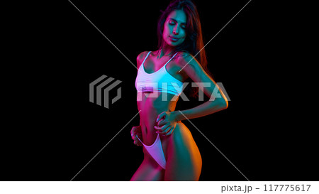 Woman posing pulling on her bikini bottom, highlighted by neon lights, showcasing her toned physique against black background. 117775617