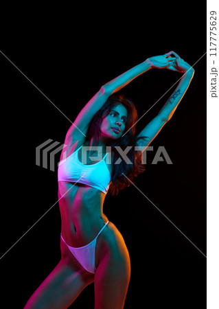 Woman stretches with grace, highlighted by vibrant neon lighting against dark background, exuding confidence and elegance. Woman stretches with grace, highlighted by vibrant neon lighting against dark background, exuding confidence and elegance. 117775629