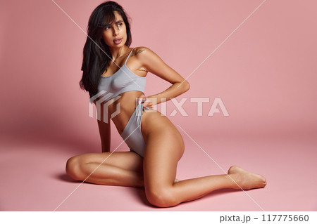 Woman kneels on floor, gracefully pulling at her lingerie, showcasing her fit physique and calm expression against soft pink background. Woman kneels on floor, gracefully pulling at her lingerie, showcasing her fit physique and calm expression against soft pink background. 117775660