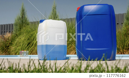 Two large chemical containers are placed poolside with a grass-level view, emphasizing the importance of pool maintenance. Two large chemical containers are placed poolside with a grass-level view, emphasizing the importance of pool maintenance. 117776162