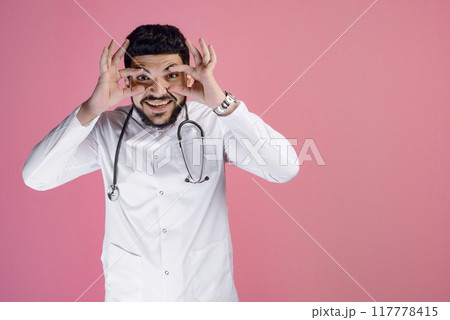 Front view of handsome male doctor wearing medical gown, looking at camera, smiling, touching eyes. Isolated on pink studio background. 117778415
