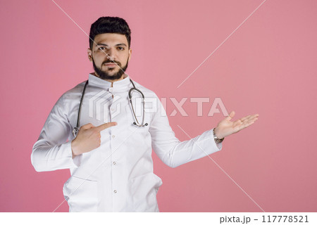Front view of young, male doctor, intern, pharmacist standing, posing indoors, pointing by fingers. Isolated on pink studio background. 117778521