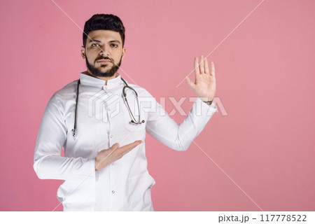 Handsome male doctor, intern standing, looking at camera, raising hand, showing. Isolated on pink studio background. 117778522