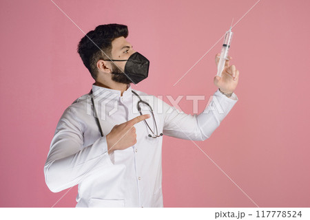 Side view of young doctor, intern wearing mask and uniform with phonendoscope, pointing to syringe. Isolated on pink studio background. Side view of young doctor, intern wearing mask and uniform with phonendoscope, pointing to syringe. Isolated on pink studio background. 117778524