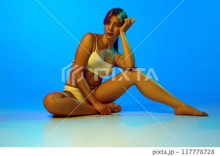 Attractive young woman with skinny body shape posing in white cotton underwear against blue studio background in neon light 117778728
