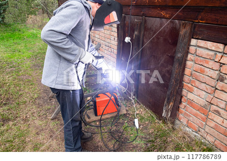 A man wearing a protective mask performs welding work outdoors near a house. Electric welding A man wearing a protective mask performs welding work outdoors near a house. Electric welding 117786789