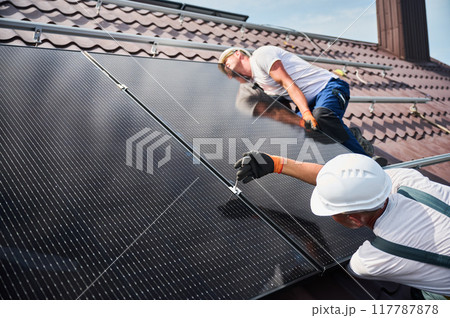 Workers building photovoltaic solar panel system on rooftop of house. Close up of men technicians in helmets and gloves installing solar module with help of hex key outdoors. Renewable energy. Workers building photovoltaic solar panel system on rooftop of house. Close up of men technicians in helmets and gloves installing solar module with help of hex key outdoors. Renewable energy. 117787878