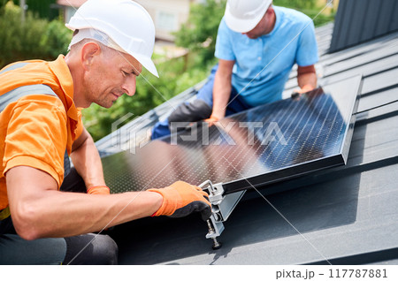 Workers building photovoltaic solar panel system on rooftop of house. Men technicians in helmets and gloves installing solar module with help of hex key outdoors. Alternative energy generation concept 117787881
