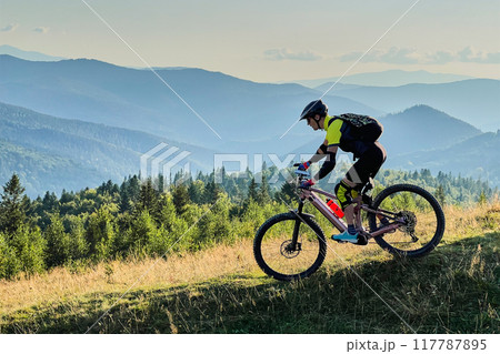 Cyclist man riding electric mountain bike outdoors. Male tourist biking along grassy trail in the mountains, wearing helmet and backpack. Concept of sport, active leisure and nature. 117787895