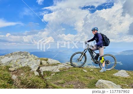 Cyclist man riding electric bike outdoors. Male tourist resting on the top of hill, enjoying beautiful mountain landscape, wearing helmet and backpack. Concept of active leisure. 117787902