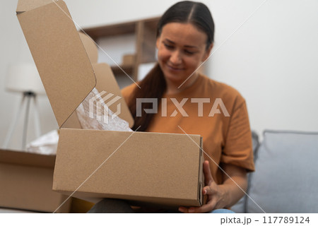 Online Bliss: Seated comfortably, a happy lady unpacks a cardboard box, expressing satisfaction with her internet purchase and express delivery. Online Bliss: Seated comfortably, a happy lady unpacks a cardboard box, expressing satisfaction with her internet purchase and express delivery. 117789124