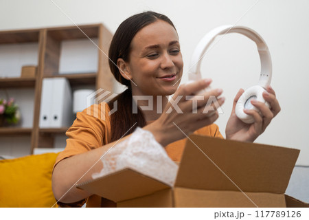 Positive woman rejoices at modern headphones from parcel delivered from online store home delivery and unpacking cardboard package with goods in housing Positive woman rejoices at modern headphones from parcel delivered from online store home delivery and unpacking cardboard package with goods in housing 117789126