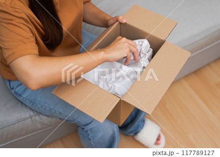 Home Delight: A content woman on her sofa unpacks her parcel, holding a cardboard box, embodying the joy of online shopping and delivery. Home Delight: A content woman on her sofa unpacks her parcel, holding a cardboard box, embodying the joy of online shopping and delivery. 117789127