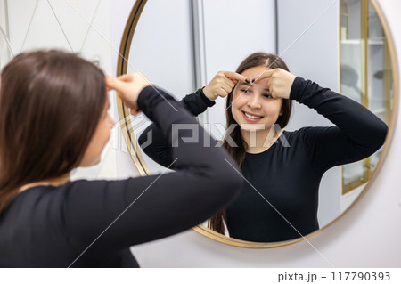 Happy young woman with long hair and a smile on her face looking in the mirror and popping a pimple on her forehead. 117790393