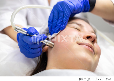 A professional dermatologist wearing medical gloves performs a microdermabrasion procedure on a smiling young woman in a professional beauty salon. 117790398