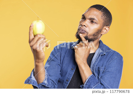 Young adult looks at an apple and thinks what can he cook with it, admiring colorful ripe fruit from local farmers market. Pensive man preparing to do a meal or dessert in studio. Young adult looks at an apple and thinks what can he cook with it, admiring colorful ripe fruit from local farmers market. Pensive man preparing to do a meal or dessert in studio. 117790678