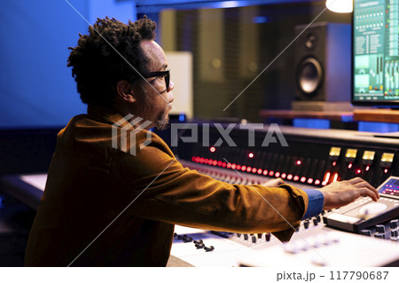 African american record producer adjusting sound settings on control board desk, editing tunes in professional studio. Music engineer working on mixing console to edit music, pushing faders. 117790687