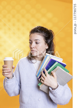 Nervous woman with troubled look on face carrying books and coffee cup, studio background. Student feels disturbed before university assessment, holding textbooks, drinking caffeine to gain courage Nervous woman with troubled look on face carrying books and coffee cup, studio background. Student feels disturbed before university assessment, holding textbooks, drinking caffeine to gain courage 117790735