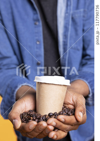 Young adult presenting cup of caffeine hot drink in front of the camera, brewing black roasted coffee beans. African american person serving cold brew beverage with delicious arabica flavor. Close up. Young adult presenting cup of caffeine hot drink in front of the camera, brewing black roasted coffee beans. African american person serving cold brew beverage with delicious arabica flavor. Close up. 117790736