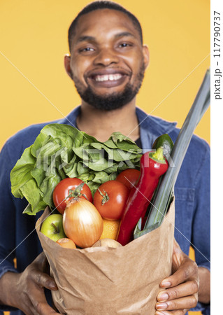 Cheerful person advocates for sustainable vegan lifestyle in studio, carrying a paper bag of fresh organic groceries. Eco friendly guy shopping to promote zero waste and delicious produce. Cheerful person advocates for sustainable vegan lifestyle in studio, carrying a paper bag of fresh organic groceries. Eco friendly guy shopping to promote zero waste and delicious produce. 117790737