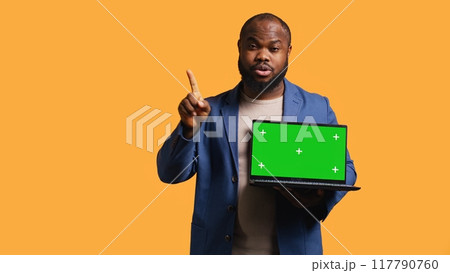 Upset african american man shows thumbs down sign gesturing holding green screen laptop, disapproving. Reviewer does rejection hand gesture regarding chroma key notebook, studio background, camera B 117790760