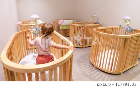 Toddler standing and playing in crib in modern nursery filled with some wooden cribs 117791338
