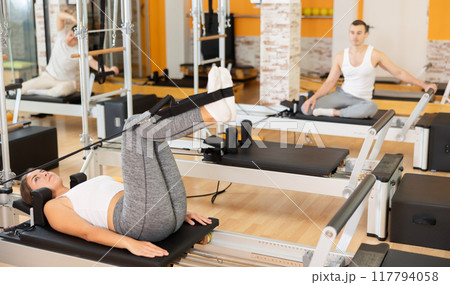 Girl works out in gym and performs set of exercises to stretch leg muscles with help of reformer 117794058