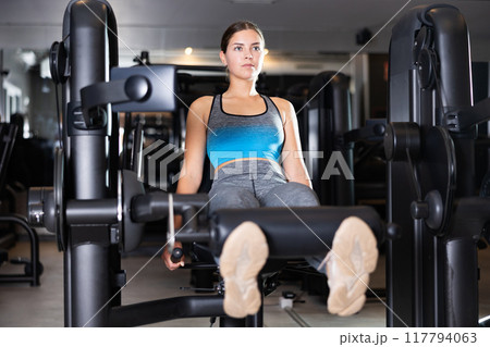 Sportive young girl training on leg extension machine in gym Sportive young girl training on leg extension machine in gym 117794063