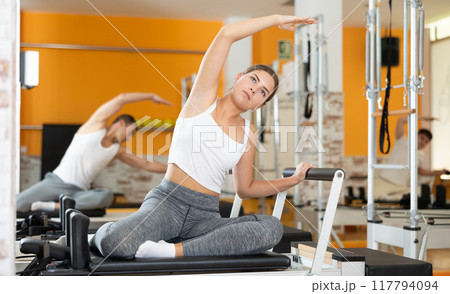 Girl sits on reformer and rests while doing exercises for muscles of body and torso. Girl sits on reformer and rests while doing exercises for muscles of body and torso. 117794094