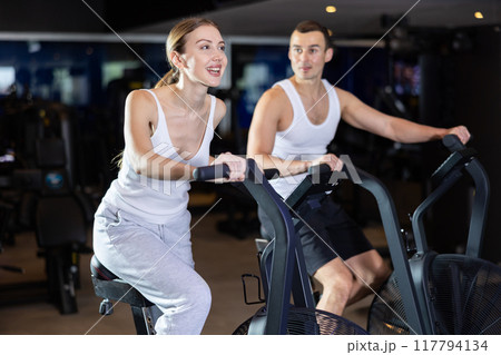 Sportive young couple doing air bike exercises in well-equipped gym Sportive young couple doing air bike exercises in well-equipped gym 117794134