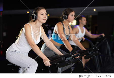 Girl in earphone performs exercises with stationary bicycle in gym Girl in earphone performs exercises with stationary bicycle in gym 117794385
