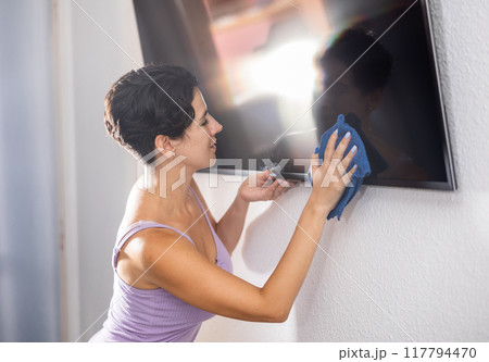 Hardworking woman wipes dust from TV with rag, tidying up apartment 117794470