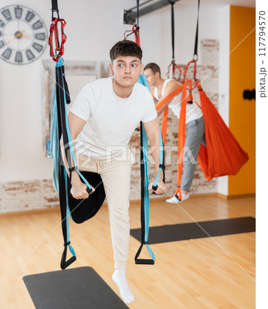 Man visitor performs exercises in hanging position on Pilates slings hammock Man visitor performs exercises in hanging position on Pilates slings hammock 117794570