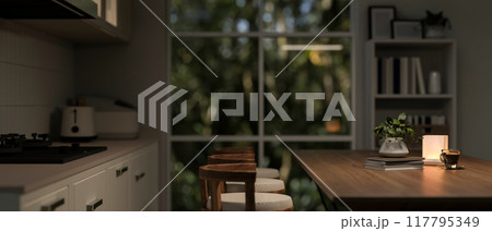 A side view image of a cozy, nighttime kitchen scene featuring a wooden dining table with stools. A side view image of a cozy, nighttime kitchen scene featuring a wooden dining table with stools. 117795349