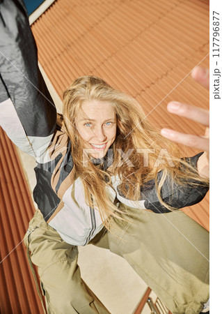 Blonde woman with blue eyes smiling joyfully while climbing on metal structure against rust-colored backdrop 117796877