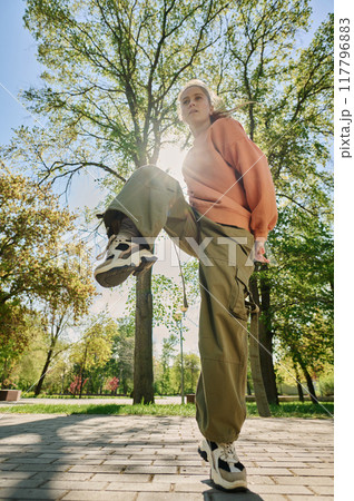 Young woman performing an outdoor exercise in a sunlit park with green trees and pathways around wearing comfortable casual clothing enjoying a sunny day Young woman performing an outdoor exercise in a sunlit park with green trees and pathways around wearing comfortable casual clothing enjoying a sunny day 117796883