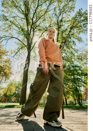 Smiling woman enjoying peaceful park environment surrounded by tall trees and green foliage while standing on paved walkway Smiling woman enjoying peaceful park environment surrounded by tall trees and green foliage while standing on paved walkway 117796885