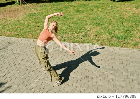 Woman performing dance move in park while casting shadow on ground, embracing lively green surroundings without natural light depicted 117796889